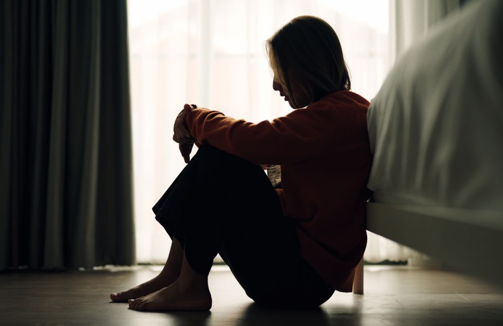 Eine Frau sitzt zusammengekauert auf dem Boden vor ihrem Bett.