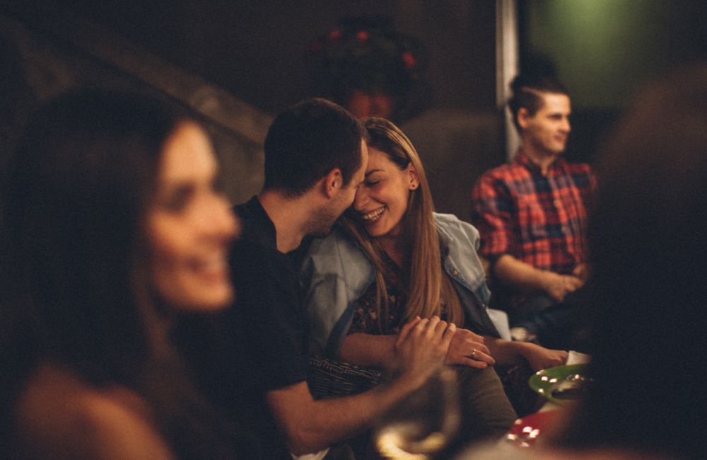 Ein Mann und eine Frau sitzen eng aneinandergeschmiegt in einer Bar und lächeln sich an.