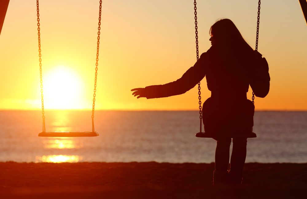 Das Bild zeigt eine Silhouette einer Person, die auf einer Schaukel sitzt, während die Sonne am Horizont über dem Meer untergeht.