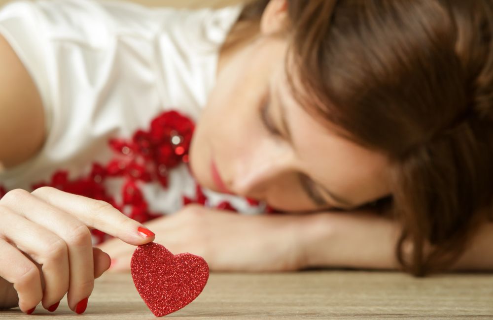 Eine Frau liegt mit ihrem Kopf auf einem Holztisch und hält mit ihrem Finger ein rotes glitzer Herz