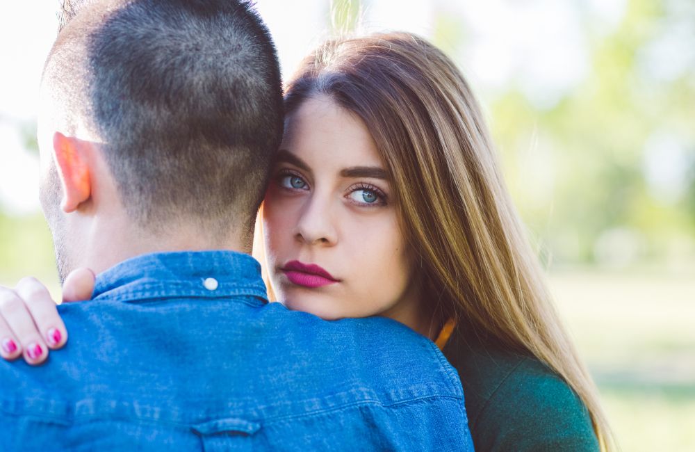 Das Bild zeigt eine Frau mit langen, braunen Haaren, die einen Mann umarmt. Ihr Gesicht ist dem Betrachter zugewandt, und ihr Blick ist nachdenklich und ernst. Der Mann ist von hinten zu sehen, trägt ein blaues Hemd und hat kurzes Haar.