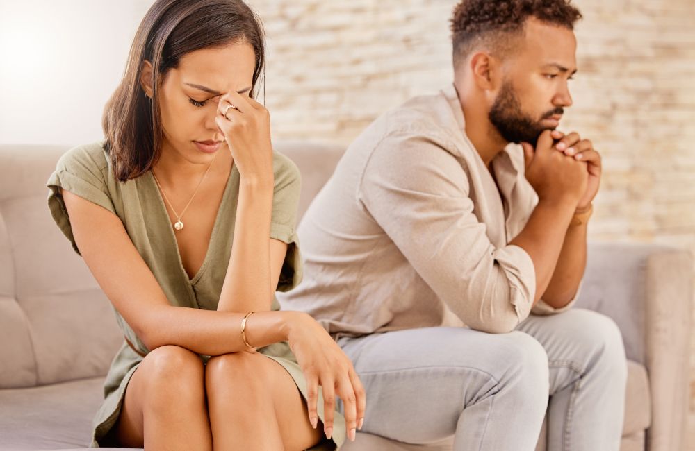 Ein Paar sitzt voneinander abgewandt auf dem Sofa, beide sehen unzufrieden und nachdenklich aus.