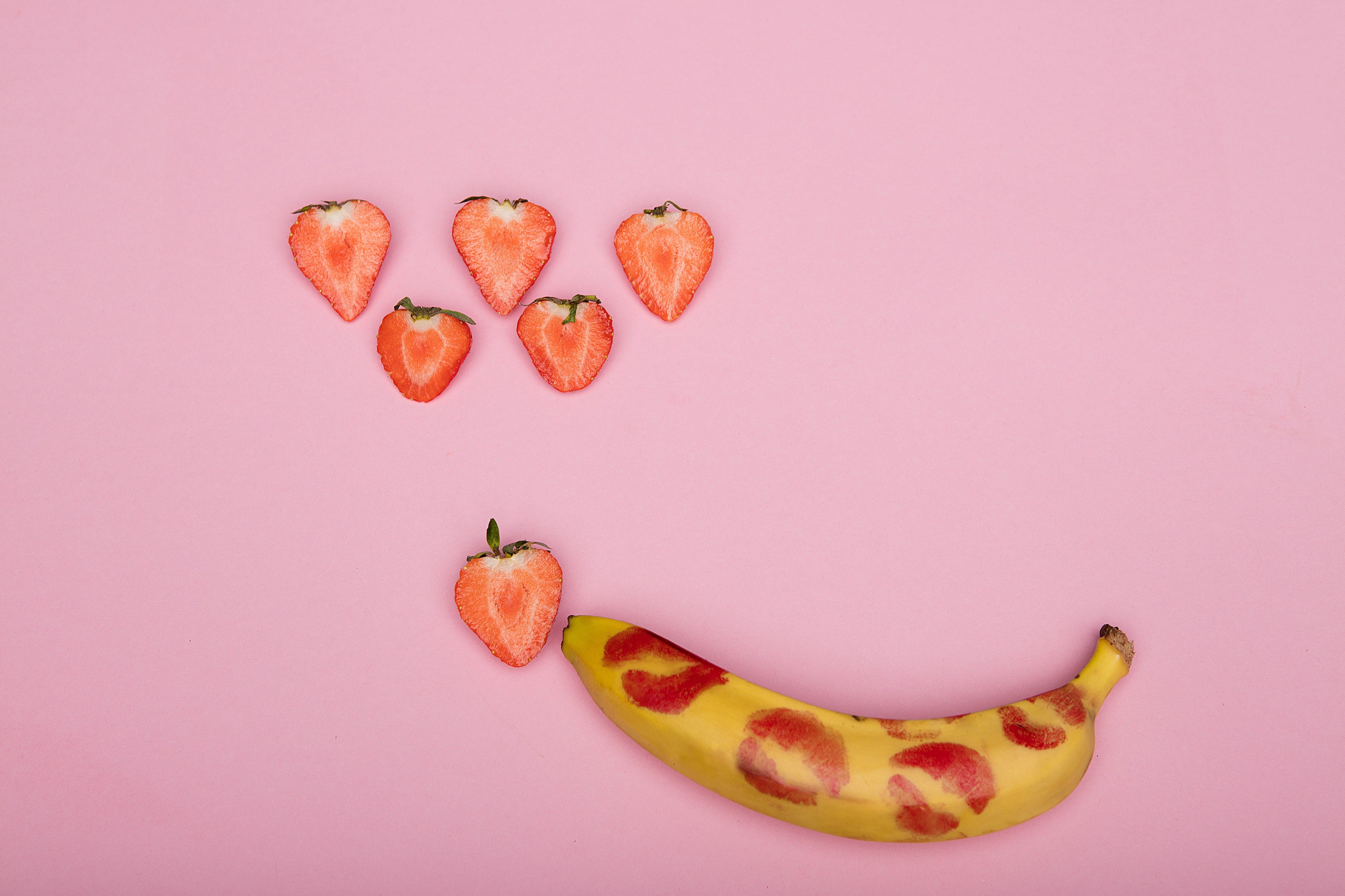 Banane mit roten Küssmündern darauf neben halben Erdbeeren auf rosa Hintergrund