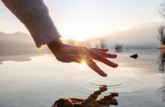 Das Bild zeigt eine künstlerische Aufnahme einer Hand, die sich über eine spiegelglatte Wasseroberfläche streckt. 