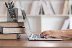 Schreibtisch mit Büchern, Stiftköcher und einem Laptop, an dem eine Person tippt vor einem Bücherregal im Hintergrund