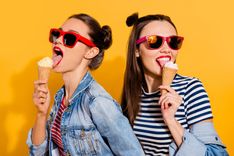 Zwei Frauen mit gestreiften Shirts, Jeansjacken und roter Sonnenbrille, die jeweils ein Eis essen vor gelbem Hintergrund