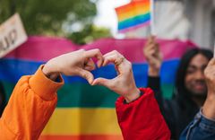 Zwei Hände formen ein Herz vor einer Regenbogenflagge