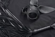 Eine BDSM Maske und ein Flogger liegen auf einem schwarten Untergrund.