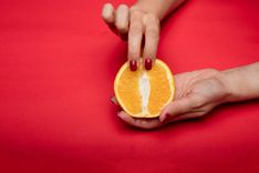 Eine Person mit roten Fingernägeln hält eine halbe Orange in der Hand und legt zwei Finger auf den oberen Teil.